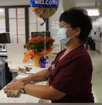 A person at a reception desk with a welcome balloon, mask on, flowers, and documents.