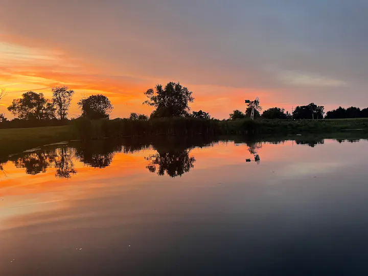 A serene sunset over a calm body of water, reflecting vibrant colors and silhouettes of trees and a windmill.