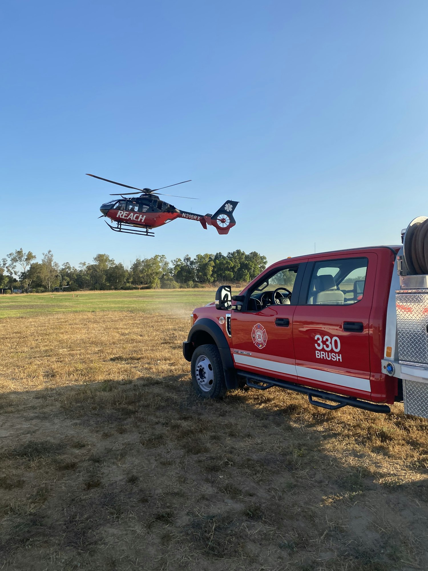 A helicopter labeled "REACH" is flying above a red brush fire truck parked in a grassy field under a clear blue sky.