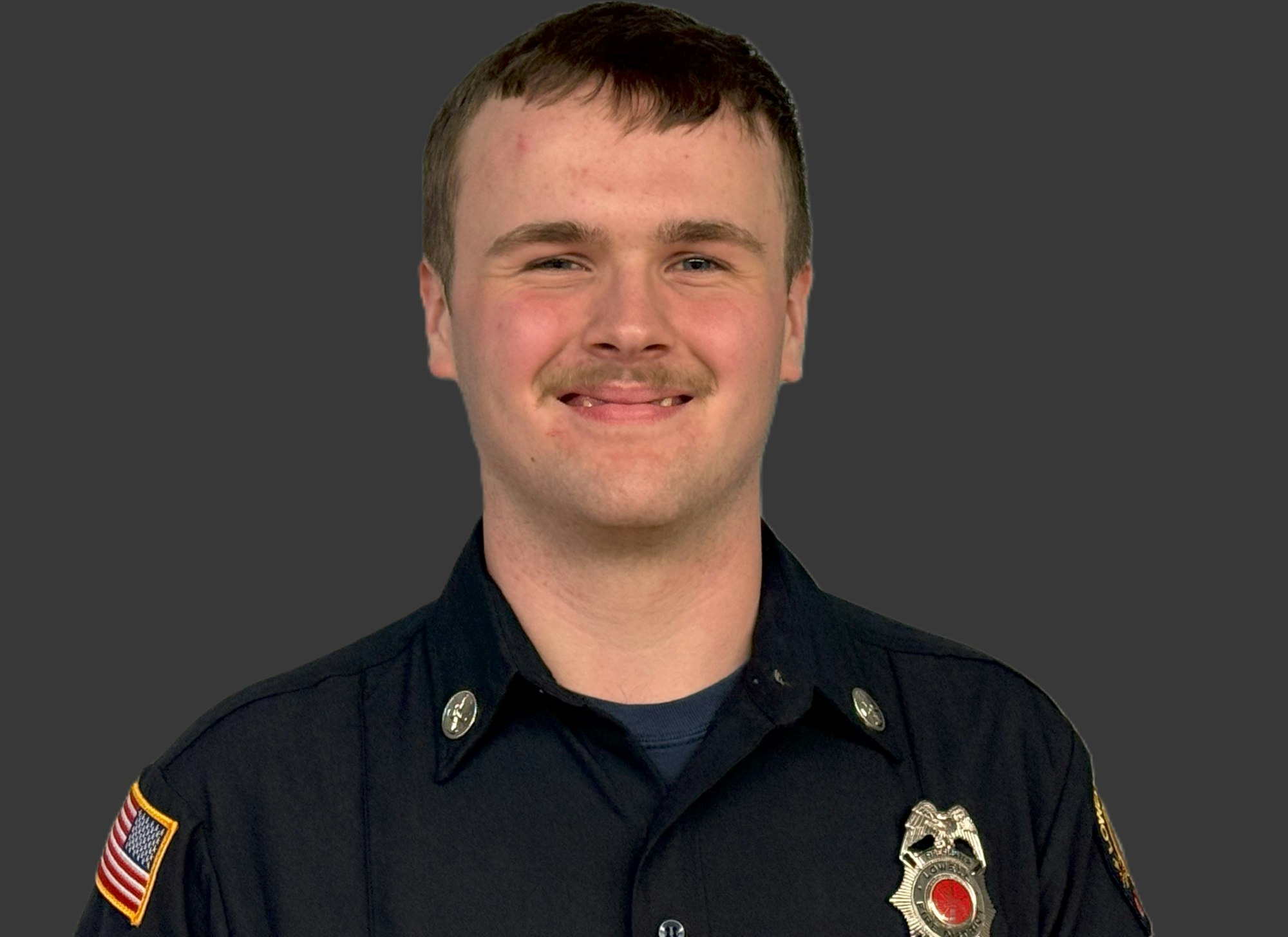 A person in a uniform with badges and a patch featuring the American flag, set against a gray background.