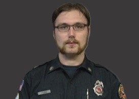 A man in a dark uniform with glasses stands against a dark background. Badges and patches are visible on his shirt.