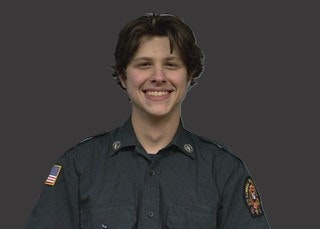 A person smiling in a uniform with patches on both sleeves, on a dark background.