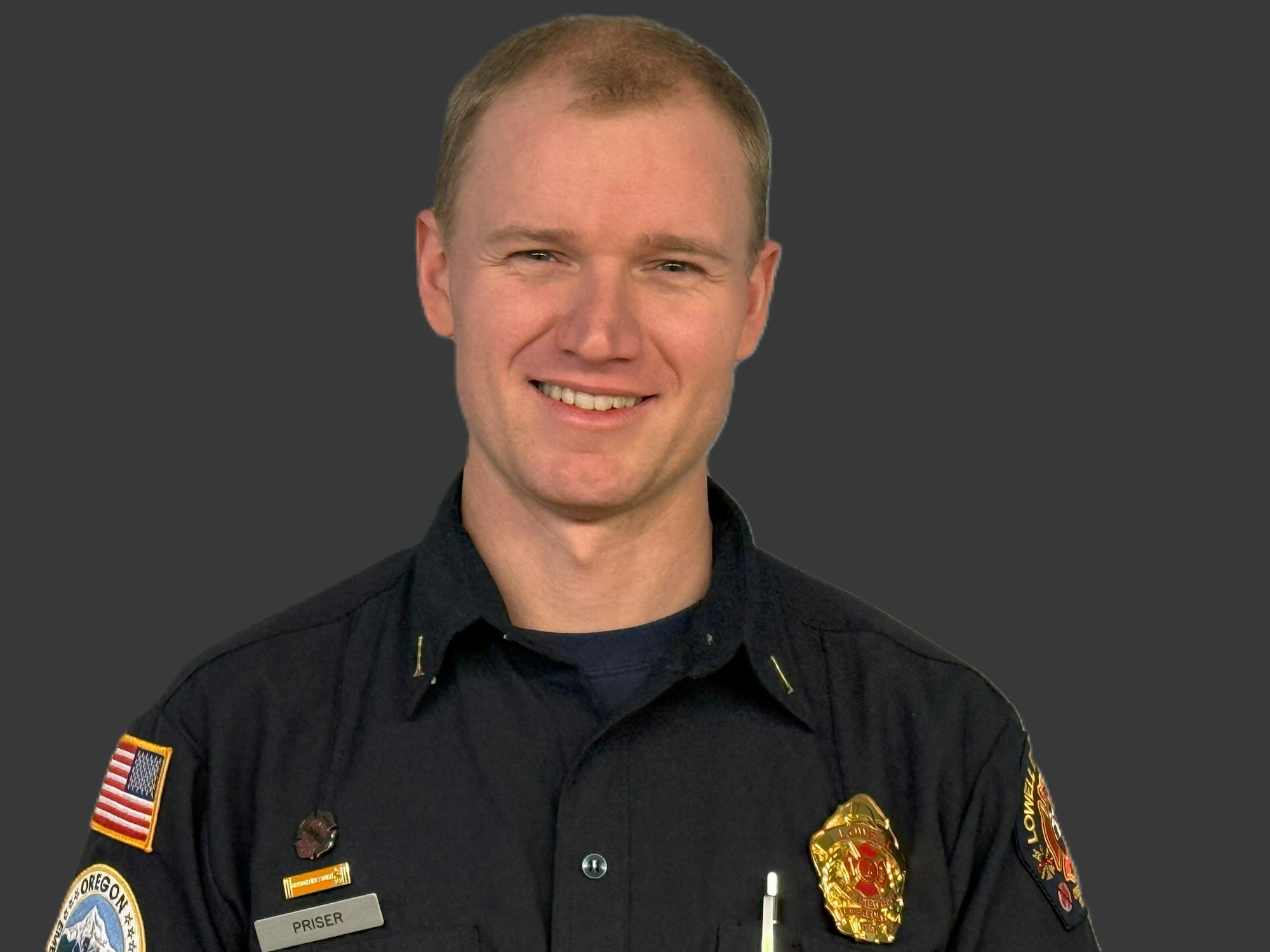 A man in uniform with patches and a badge smiling against a gray background.