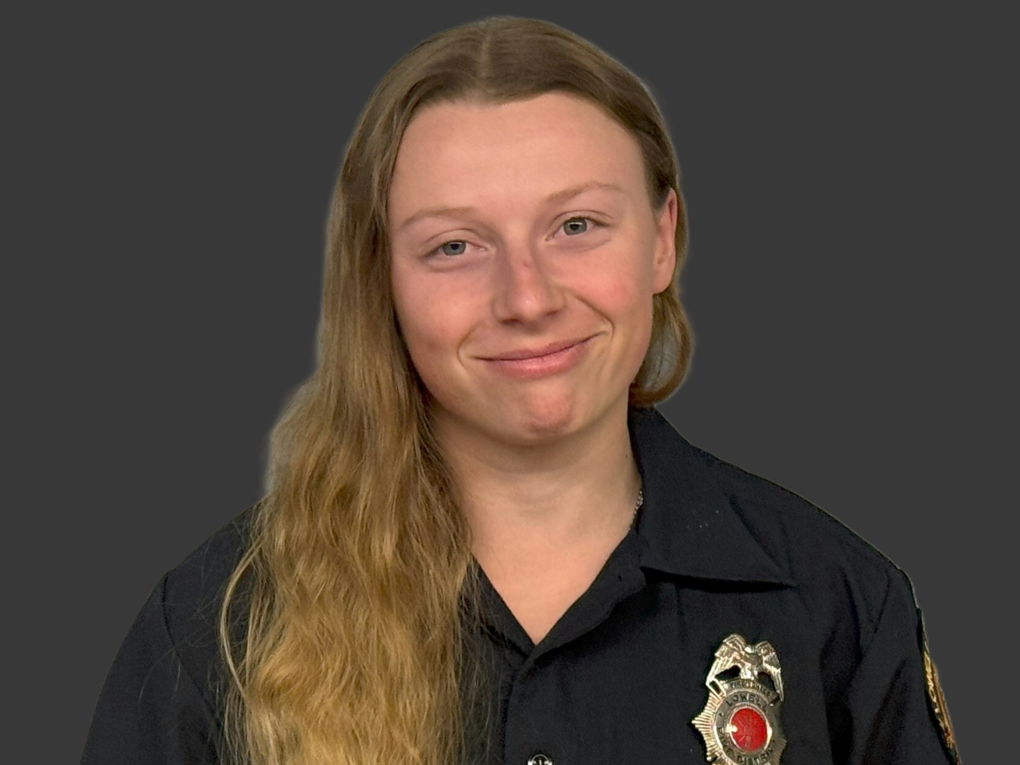 A person in a black uniform shirt with a badge, smiling against a gray background.