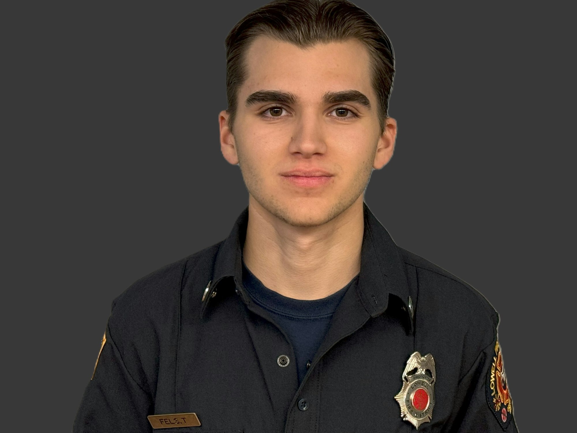 A person in a dark uniform with a badge and shoulder patch stands against a grey background.