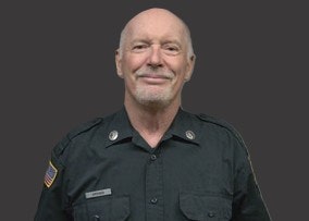 An elderly person in a dark uniform with an American flag patch on the shoulder, smiling at the camera.