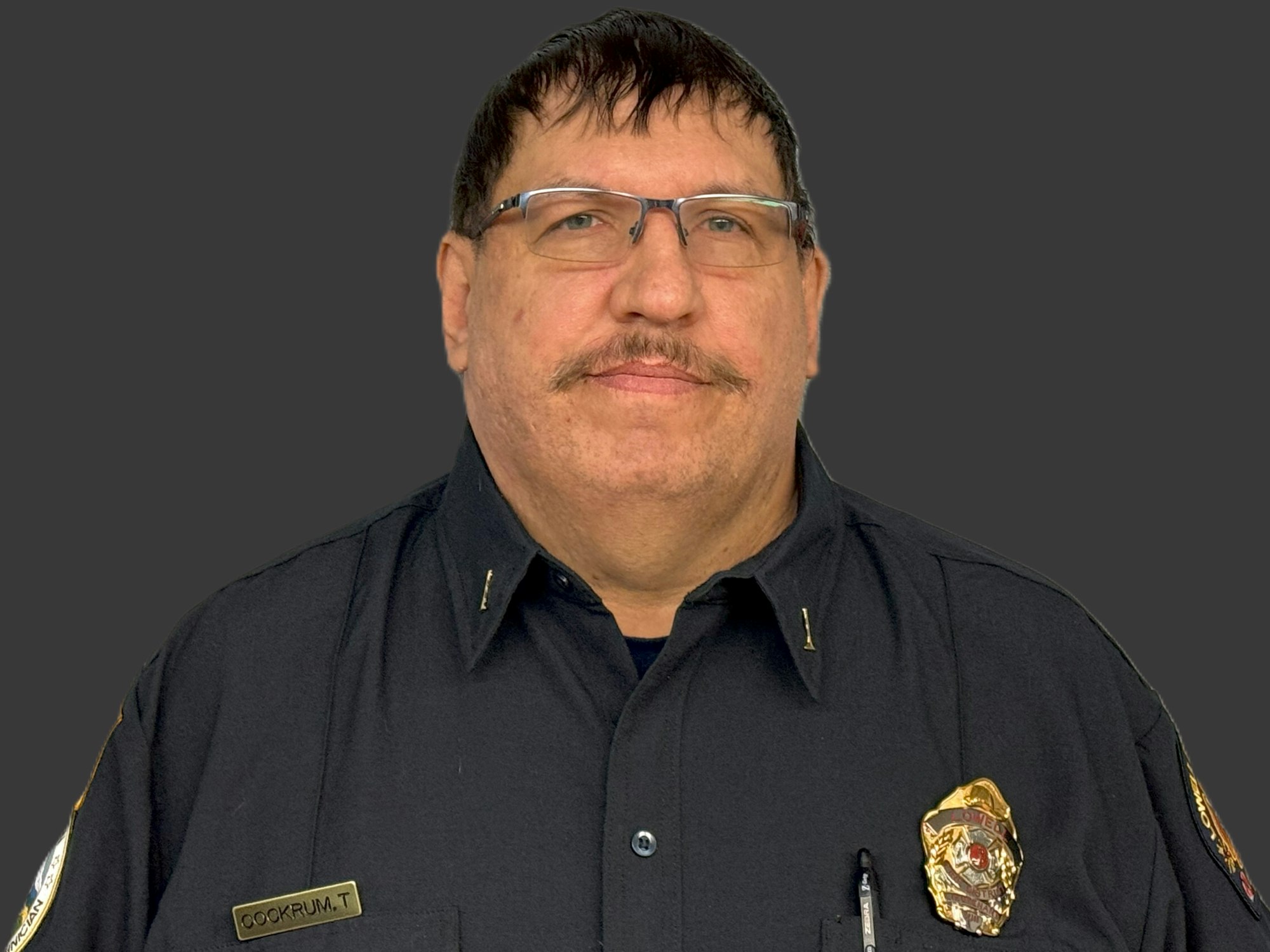A person in a uniform with a name tag and badge, against a dark background.