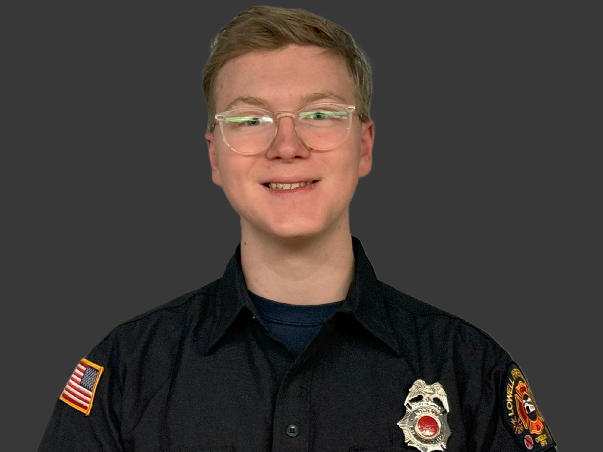 A person in a black uniform with a fire department patch and a badge, wearing glasses, against a gray background.