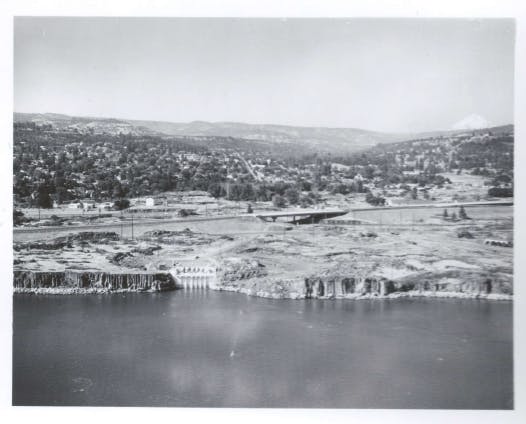 A black-and-white landscape showing a river, land with structures, and mountains in the background.