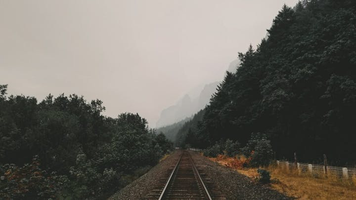 Railroad track flanked by dense trees, leading into misty mountains in the distance.