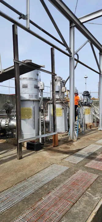 The image shows an industrial setting with machinery, piping, and a worker in an orange shirt, possibly at a manufacturing or processing facility.
