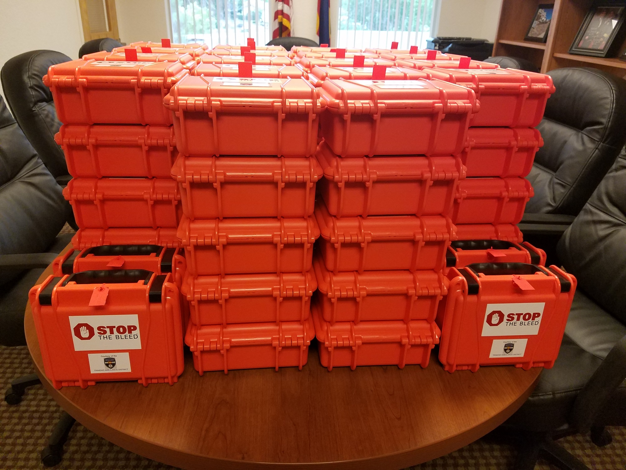 Stacks of orange "Stop the Bleed" emergency cases on a table in an office setting.