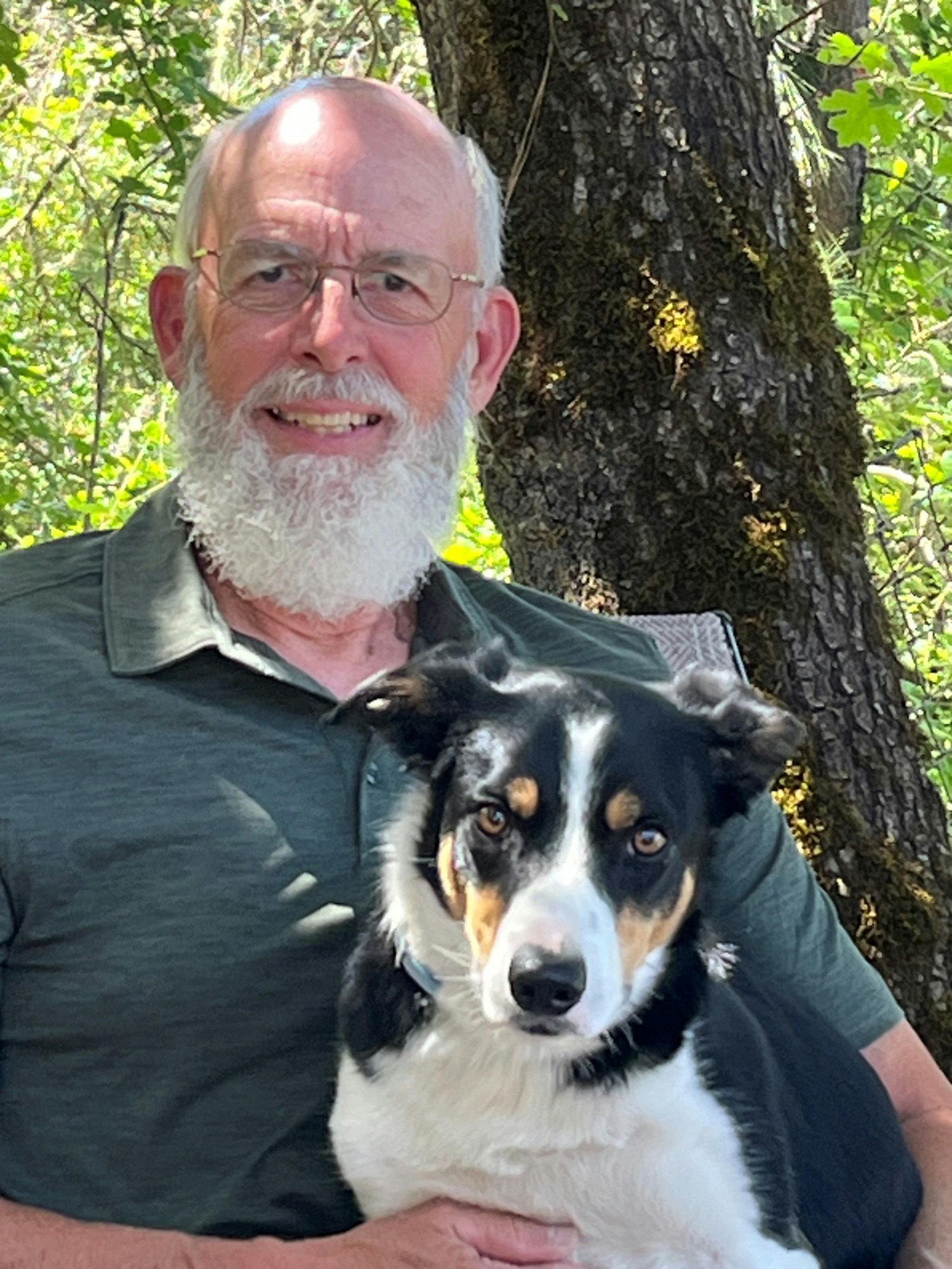 A man with a beard and glasses posing with a black and white dog outdoors.