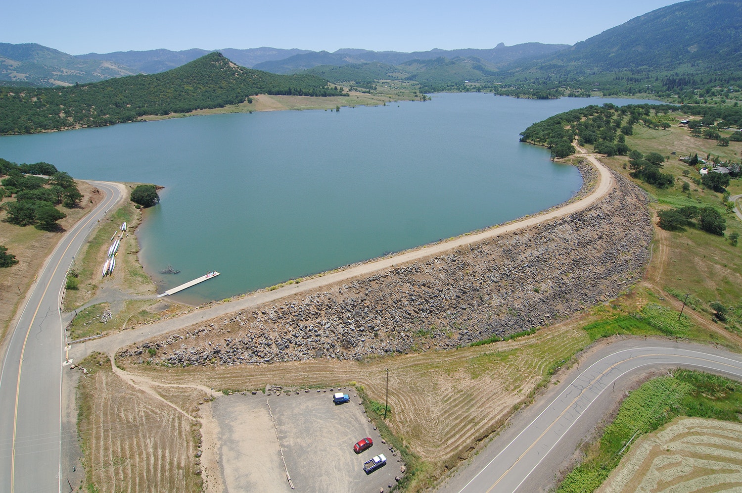 Aerial view of a serene lake surrounded by hills, with a road, a parking area, and a small dock for boats.