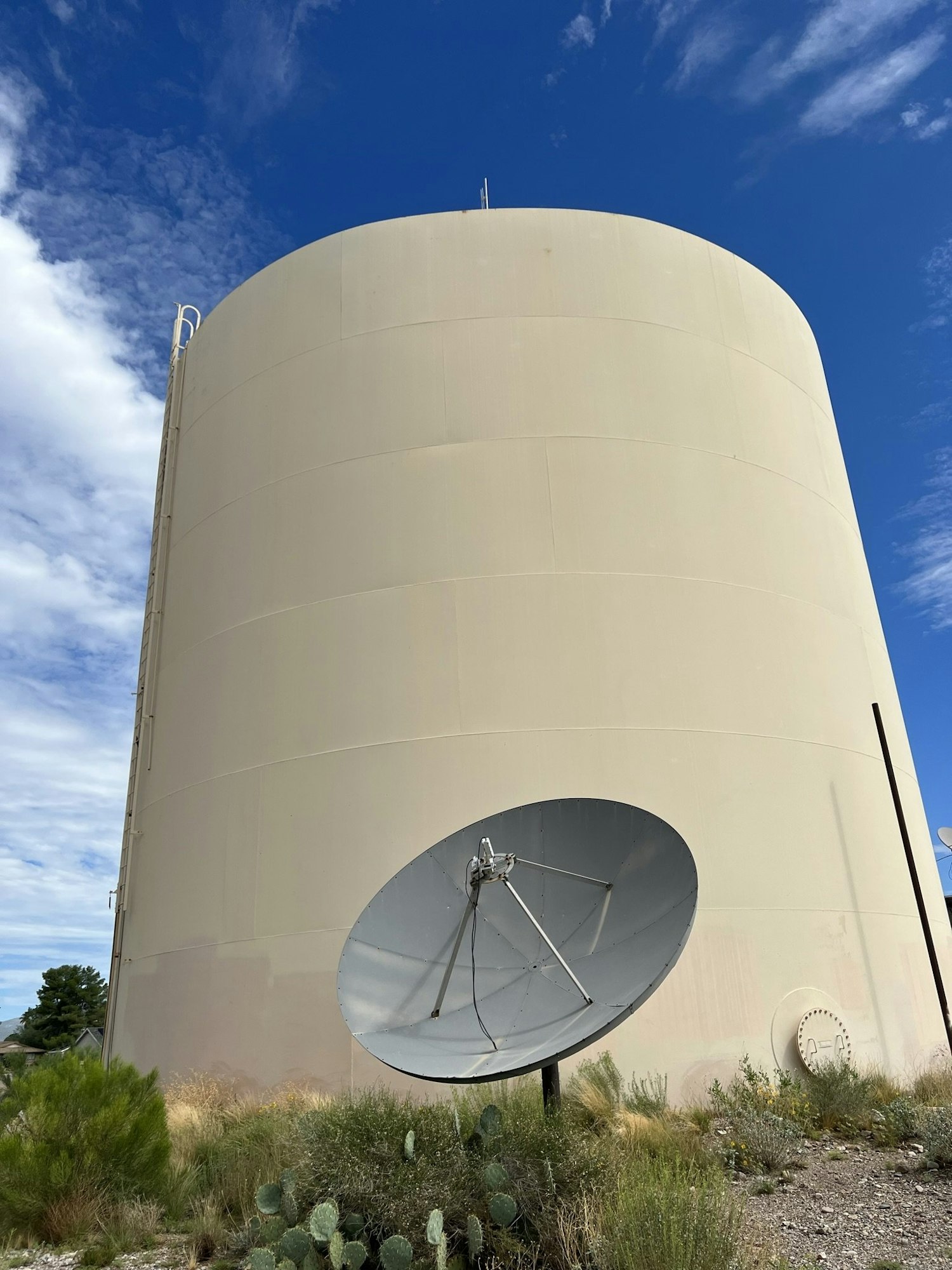 Large cylindrical structure with a satellite dish in front, surrounded by vegetation under a blue sky.