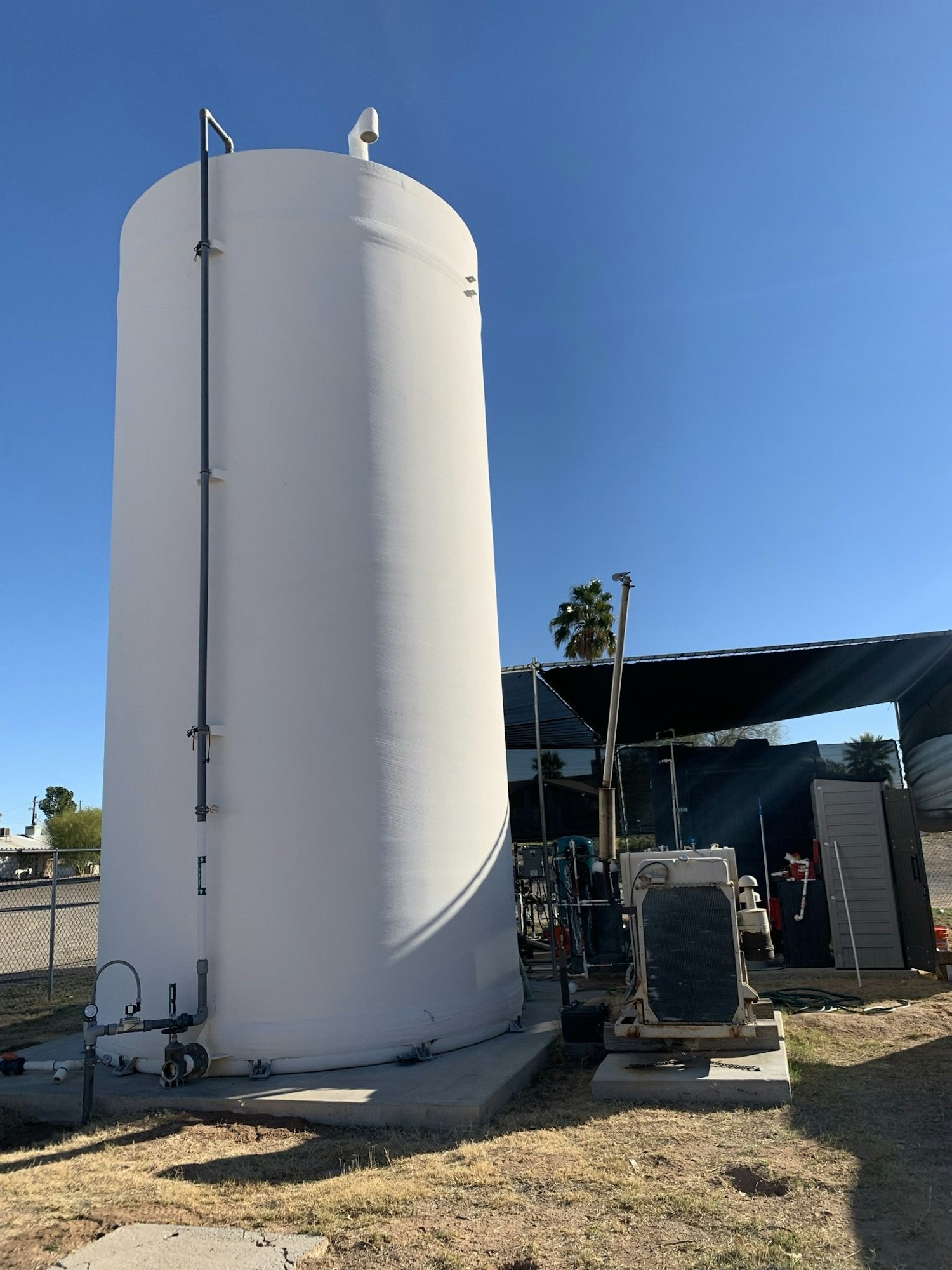 A tall white cylindrical tank with attached pipes on a concrete base, surrounded by equipment and a clear blue sky.