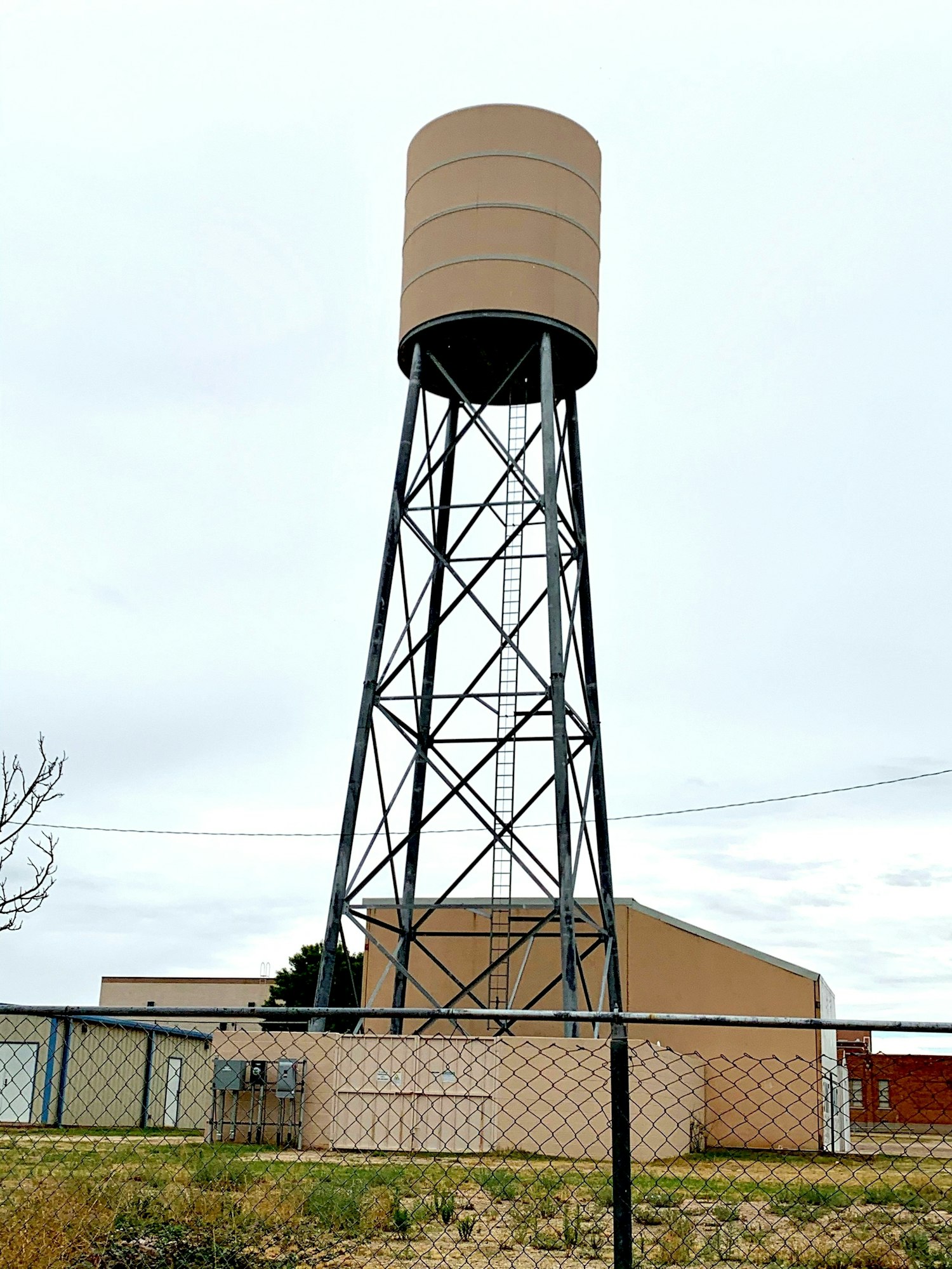 A tall water tower with a ladder, standing in front of buildings and surrounded by a chain-link fence.