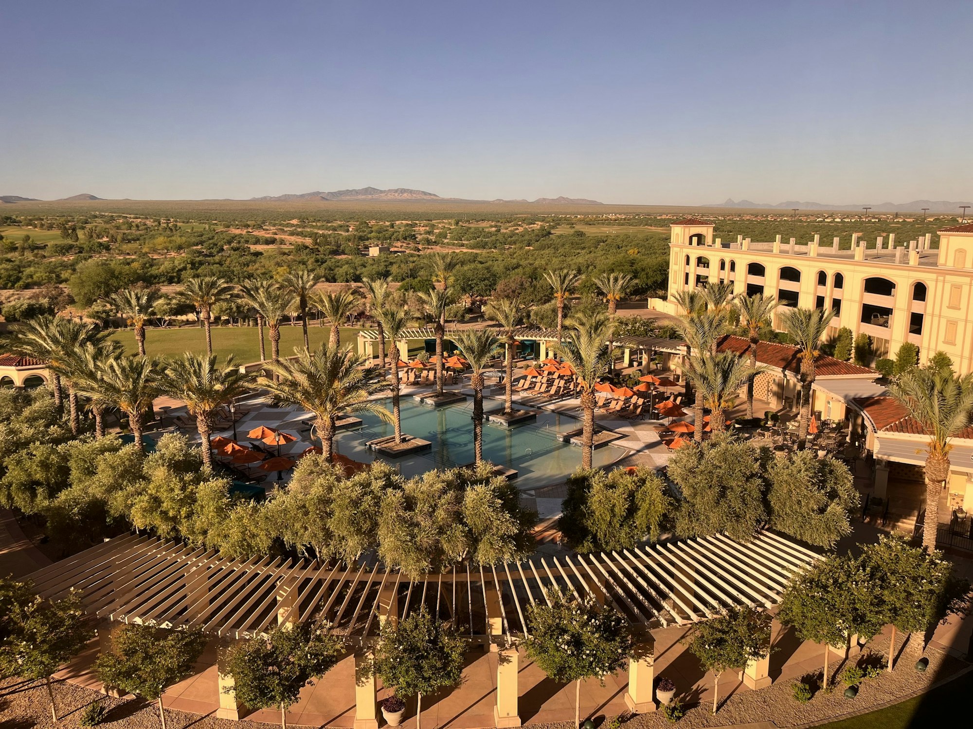 A resort with a swimming pool, palm trees, outdoor seating, and distant mountain views.