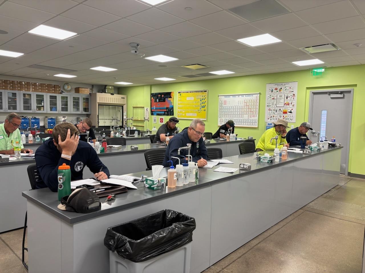 People sitting at desks in a classroom or lab, writing or studying with books and supplies on the tables.