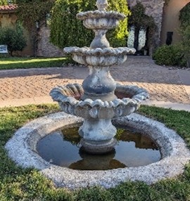 A tiered stone fountain with water, surrounded by grass and brick paths.