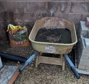 A wheelbarrow filled with water and debris, positioned between a brick wall and some stacked bricks. A bag of soil or fertilizer is nearby.