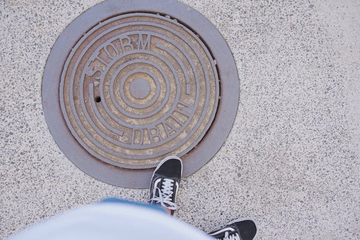 The image shows a manhole cover labeled "STORM DRAIN" and a pair of black shoes standing on a textured concrete surface.