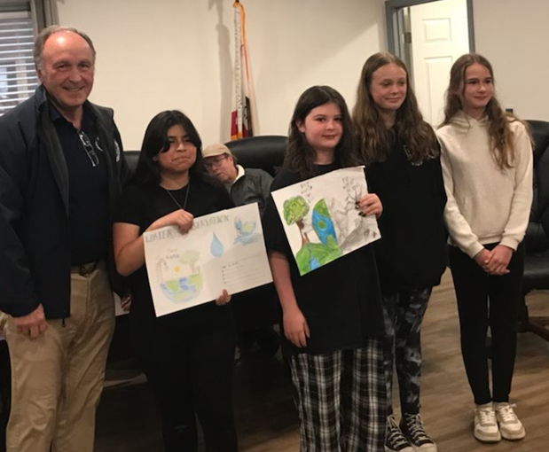A group of people, including kids holding environmental posters, pose indoors with an older man.