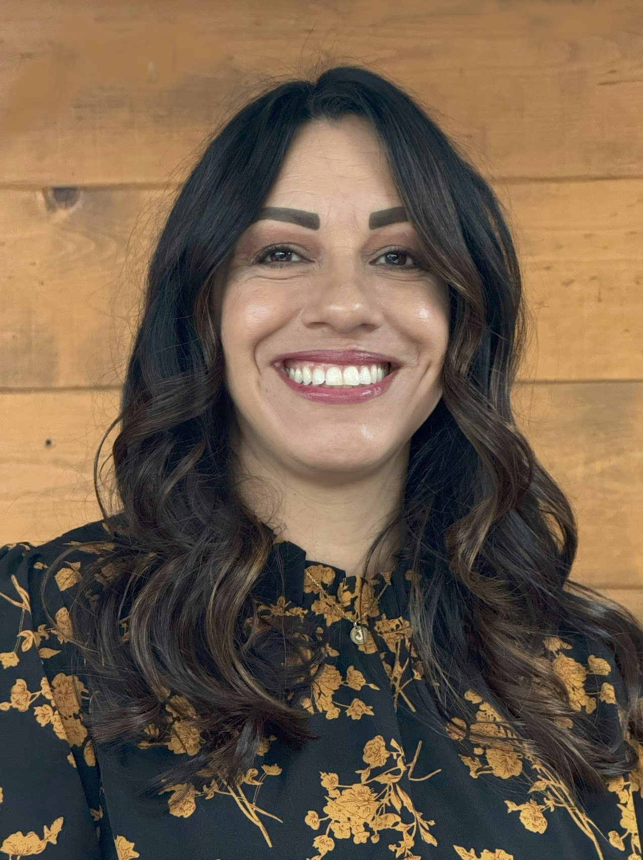 A person smiling, wearing a floral patterned shirt with a wooden background.