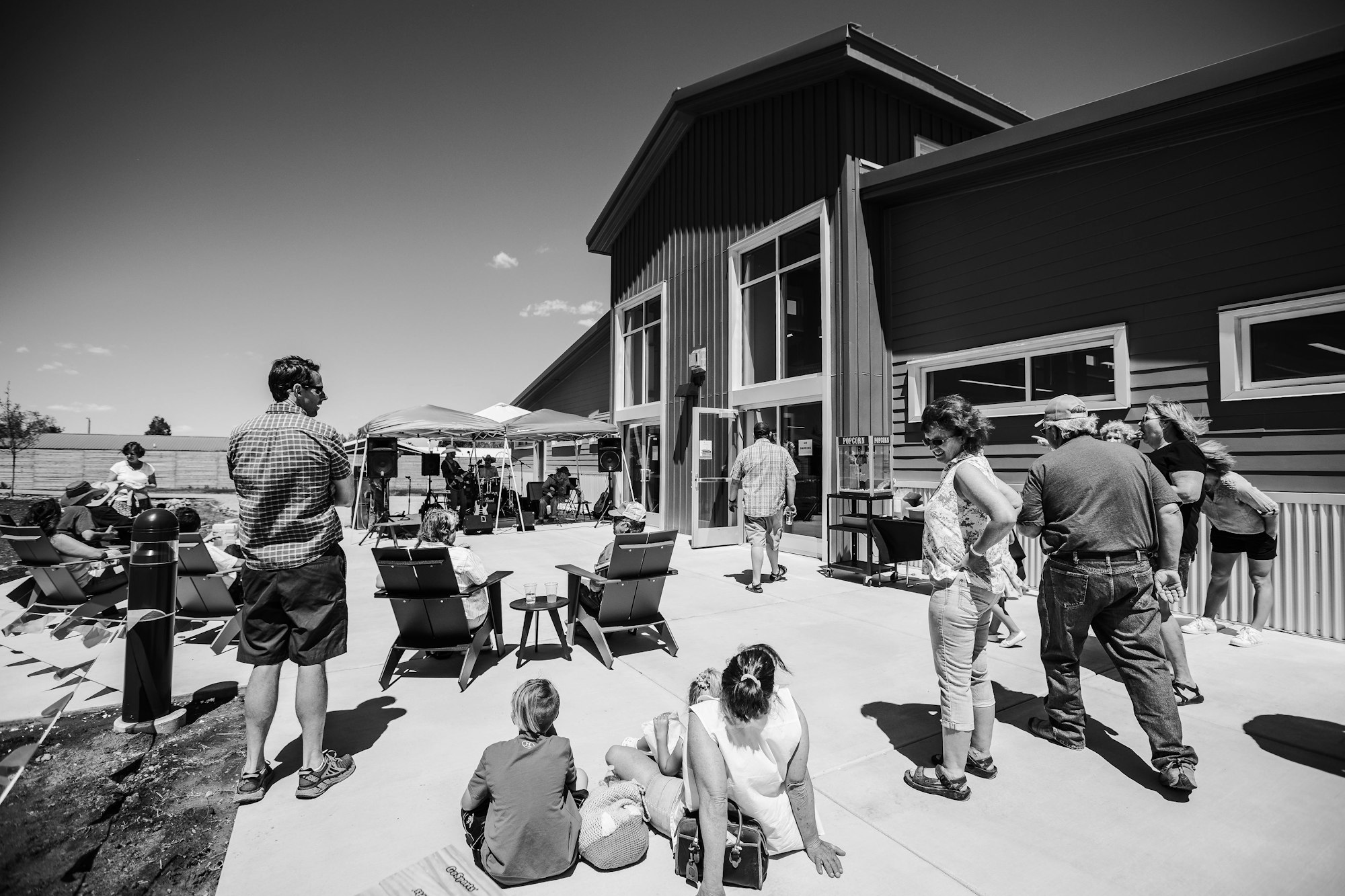 Contains people from the community outside the library for a program in the summer. Some are standing and some people are sitting with shade structures in the background.