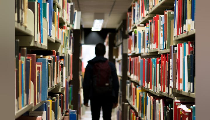 A person with a backpack walking down a library aisle lined with shelves of colorful books.
