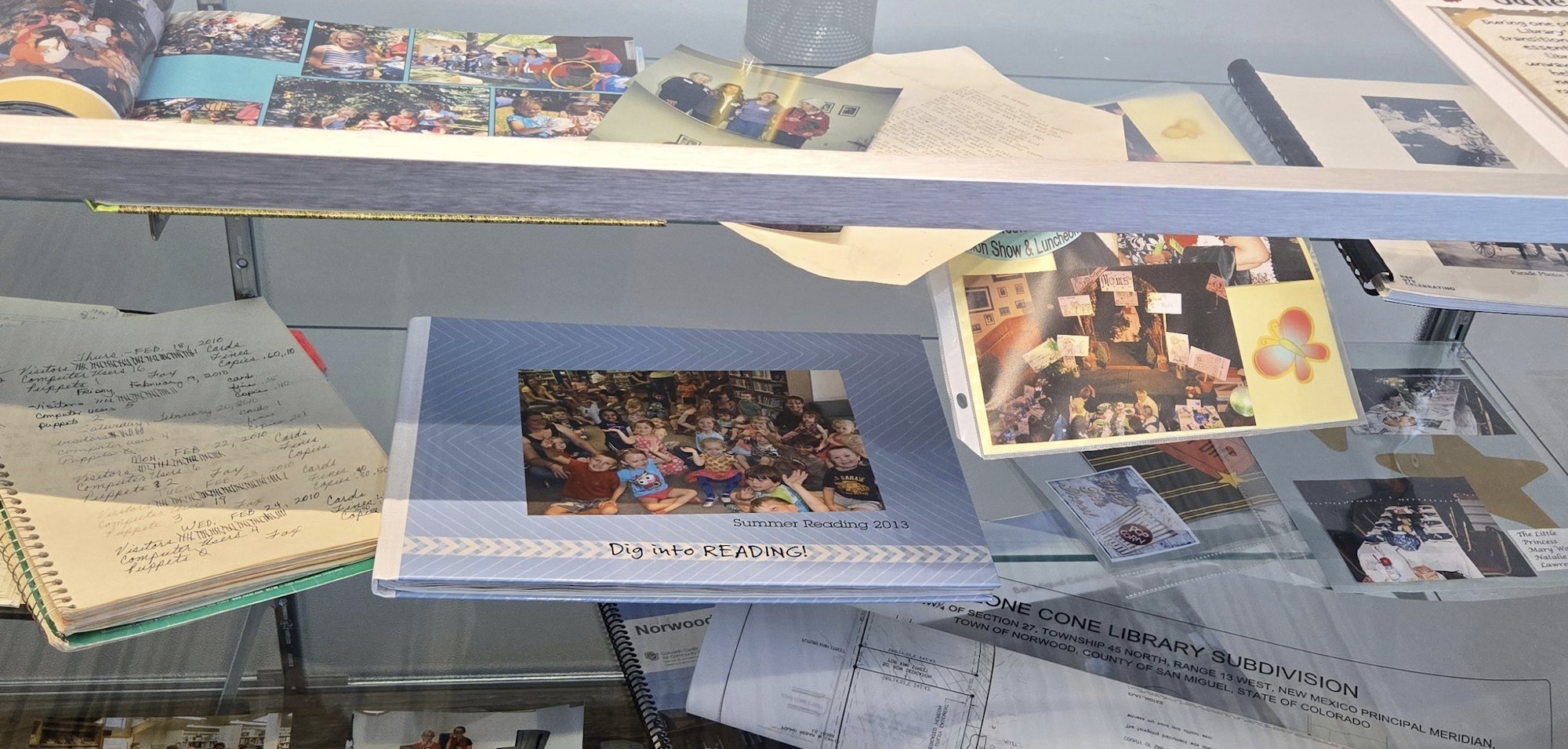 A glass display case with photos, notebooks, documents, and a “Summer Reading 2013” book.