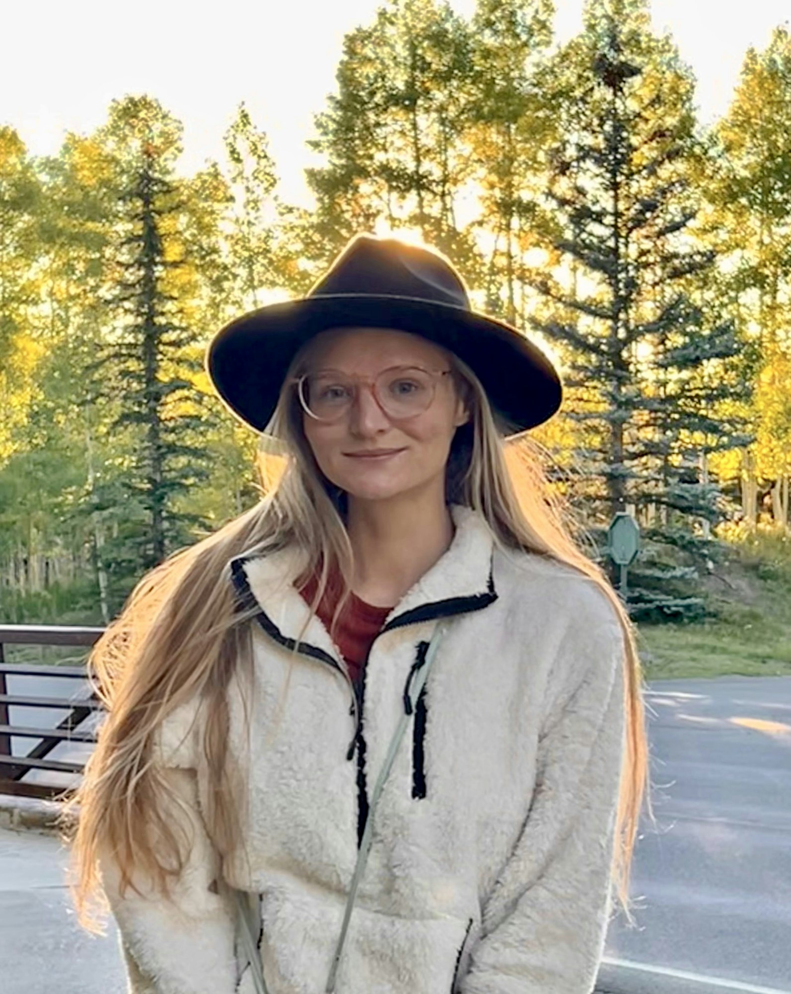 A person wearing glasses and a wide-brimmed hat, standing outdoors near trees, with sunlight filtering through the background.
