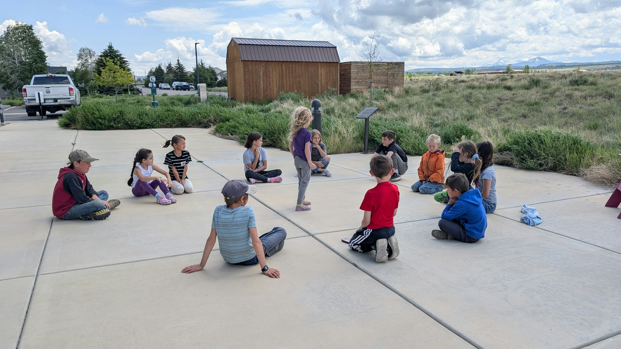 A group of children sitting in a circle outdoors, engaged in an activity near a shed and surrounded by grass and trees.