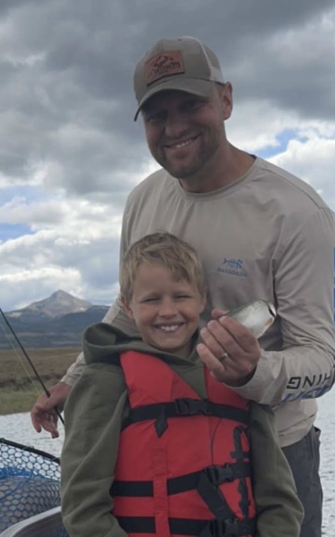 A smiling boy and a man on a boat with a fish, surrounded by Lone Cone Mountain, water and cloudy skies.