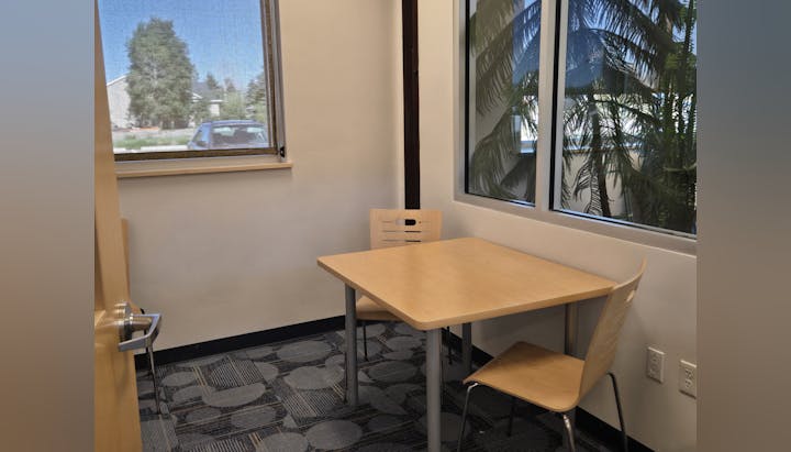 A small, empty room with one table, two chairs, patterned carpet, and a window showing trees and a parked car.