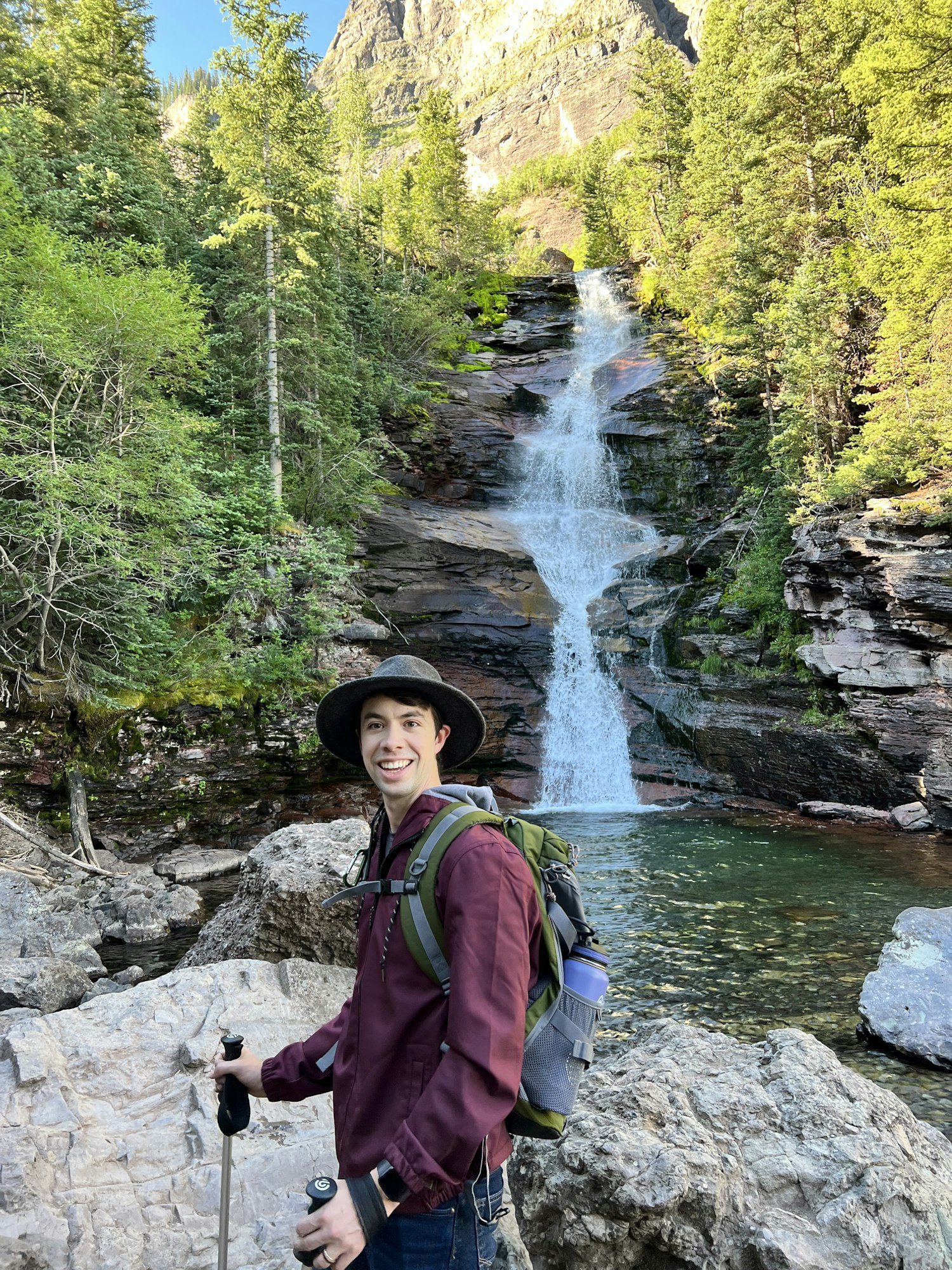 A person with a backpack and trekking poles standing in front of a waterfall amidst a forested area.