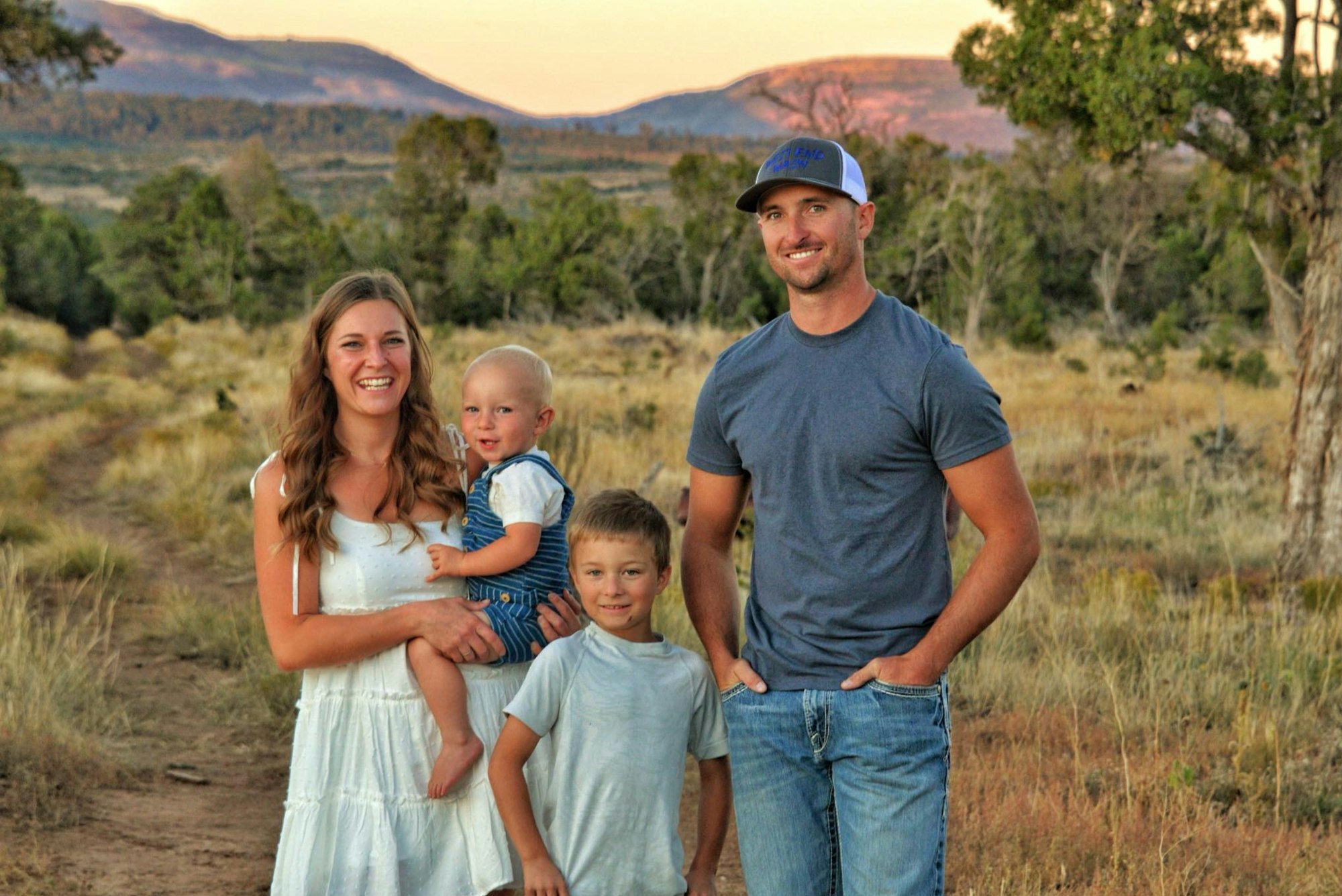 A family of four smiling outdoors in a scenic, grassy area with mountains and trees in the background.
