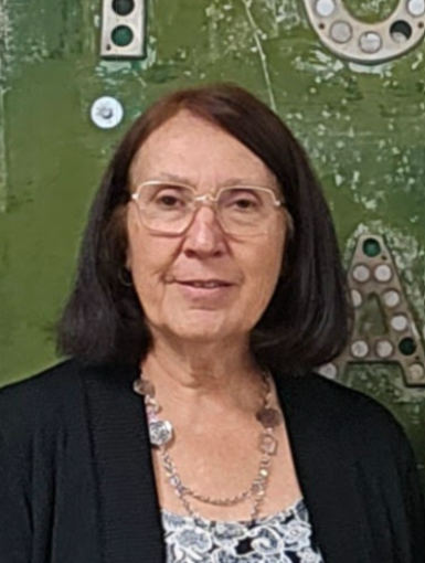 A person with glasses and dark hair, wearing a black jacket, stands in front of a green background with partially visible letters.