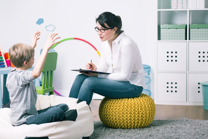 A woman with a clipboard sits on a yellow beanbag, listening to a boy with raised arms in a colorful room, possibly during a therapy session.