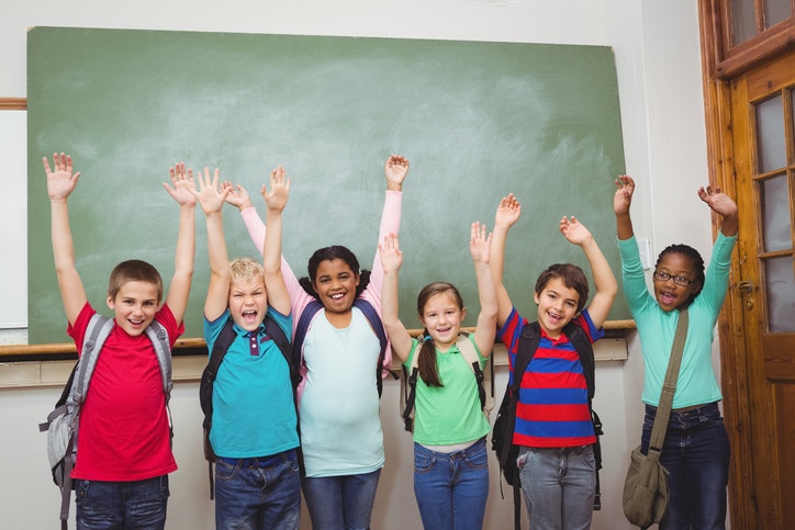 Six children with backpacks raise their arms in front of a chalkboard, smiling.