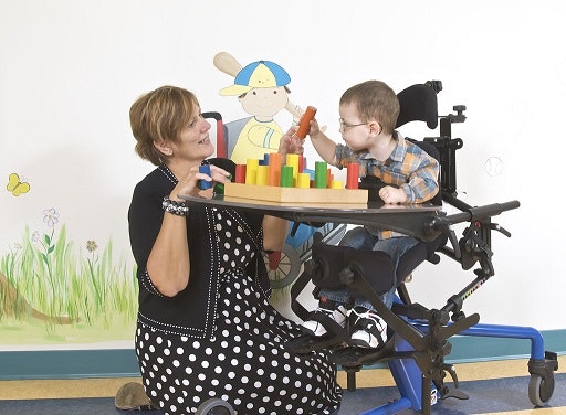 A woman interacts with a child in a wheelchair, who is playing with colorful blocks on a tray.