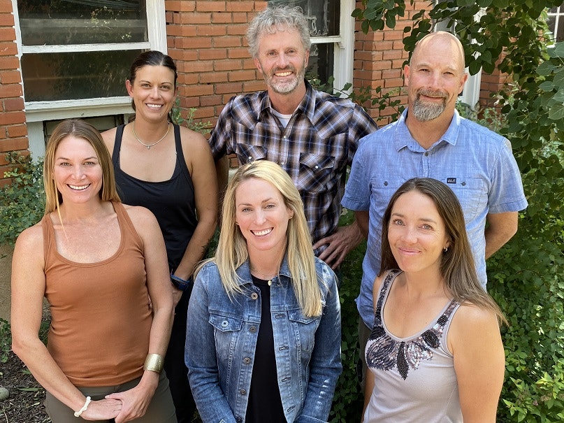 A group of six people smiling outdoors, standing in front of a brick wall with greenery.