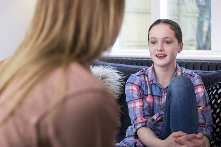 Two people sit on a couch, engaging in conversation; one person faces the other, who sits cross-legged wearing a plaid shirt.