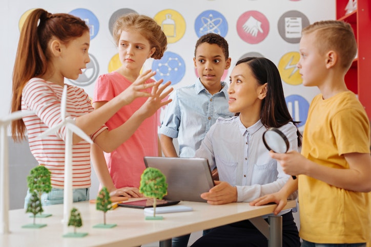 A group of kids with a teacher engaged in a science lesson, with wind turbines and trees on the table.