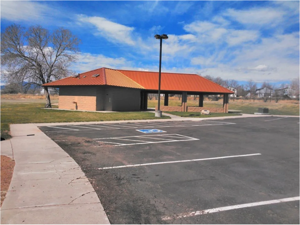 A small building with a red roof, an adjacent covered area, parking lot with handicapped space, trees, and a clear blue sky.