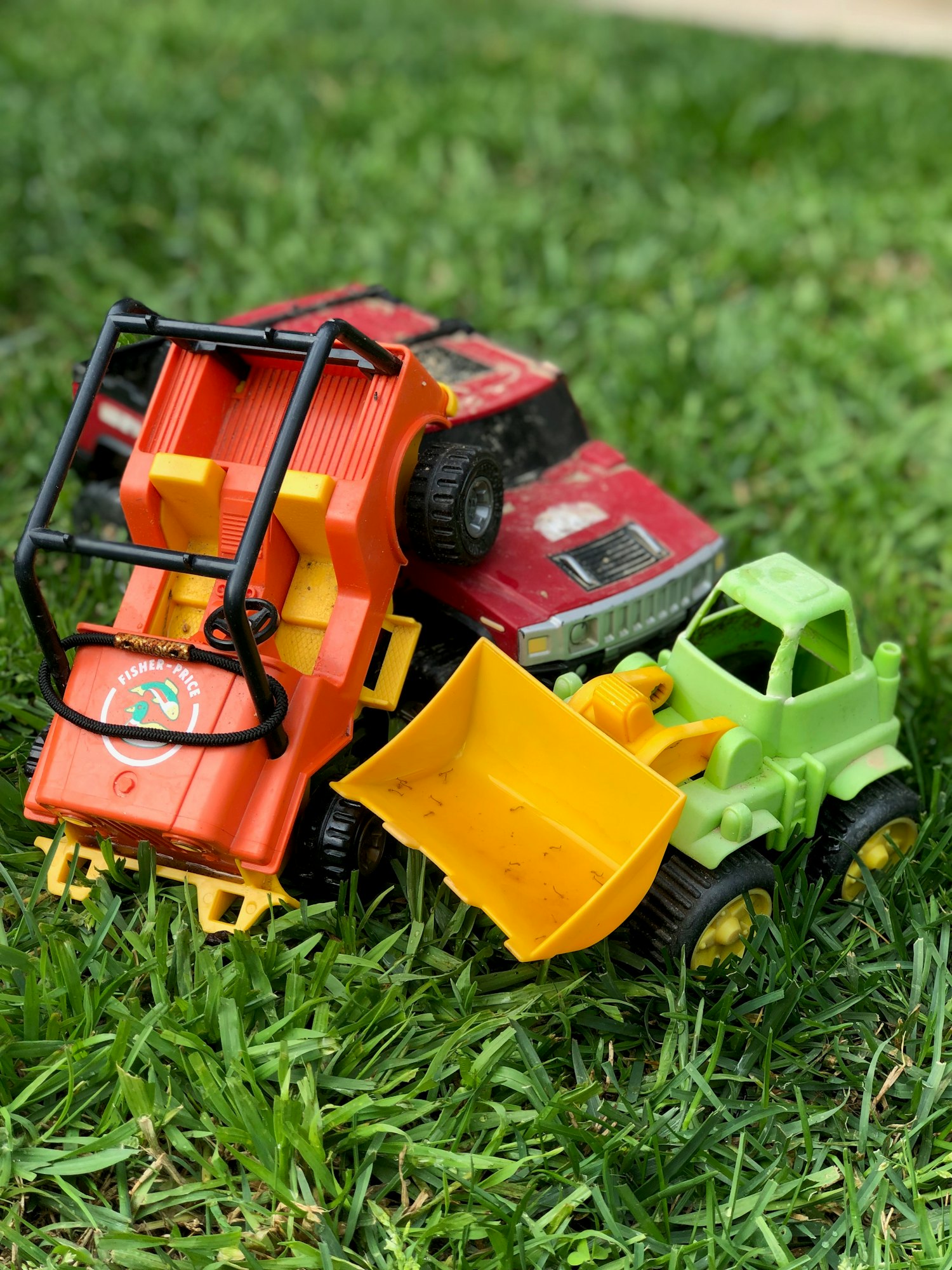 Toy trucks and a bulldozer on grass; one red, one orange with yellow, and one green with a yellow scoop.