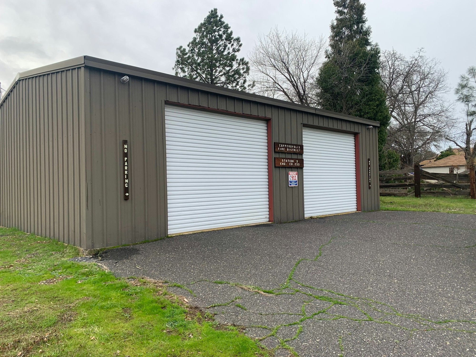 A gray building with two closed white roller doors, a "NO PARKING" sign, and some greenery around.