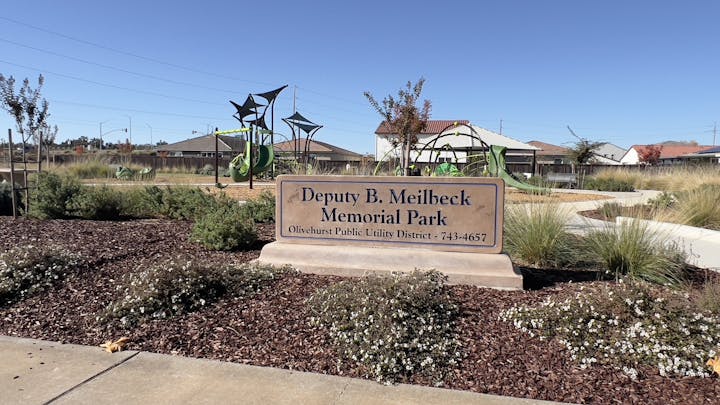 A park sign for "Deputy B. Meilbeck Memorial Park" with a landscaped area and playground structures in the background.