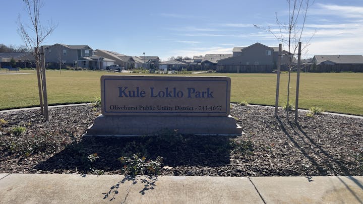 Sign reading "Kule Loklo Park" with grass, trees, and houses in the background.