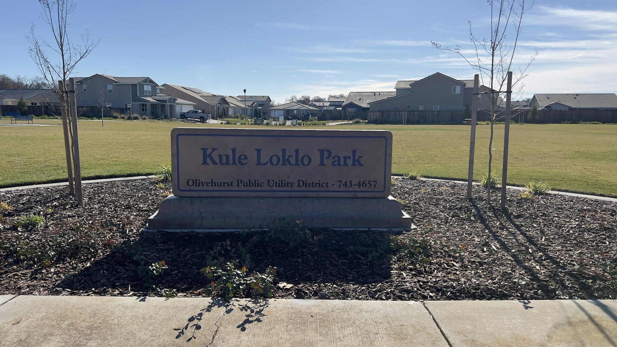 Sign reading "Kule Loklo Park" with grass, trees, and houses in the background.